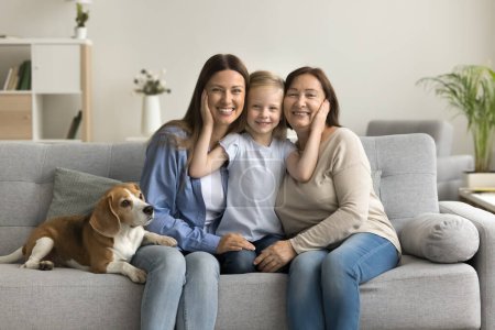 Photo for Female family portrait with adorable beagle dog. Happy mother, grandmother and little child girl sitting close on sofa, hugging with love, affection, looking at camera, smiling - Royalty Free Image