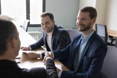 Téléchargez les photos : Heureux hommes d'affaires qui se serrent la main à la table de réunion du bureau, souriant, riant, discutant de contrat de travail, d'accord, d'accord. Chef d'entreprise, patron donnant poignée de main à l'employé avec reconnaissance - en image libre de droit