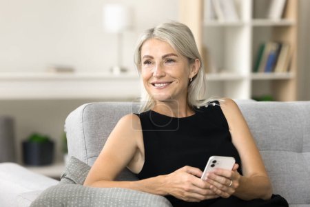 Photo for Happy dreamy middle aged woman holding smartphone, thinking on application for Internet communication, looking away with pensive face, dreaming, texting message, smiling - Royalty Free Image
