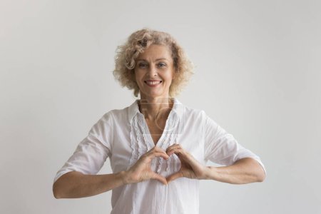 Photo for Happy pretty mature lady making heart shaped fingers, applying hands to chest, showing gesture of kindness, care, love, romance, smiling at camera, posing for portrait at white background - Royalty Free Image