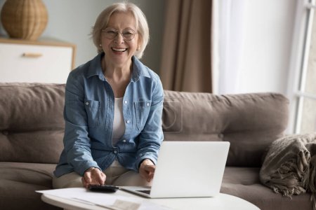 Photo for Cheerful pretty senior pensioner woman using laptop, calculator at home, looking at camera, smiling, laughing. Retired lady counting income, savings, satisfied with profit. Female portrait - Royalty Free Image