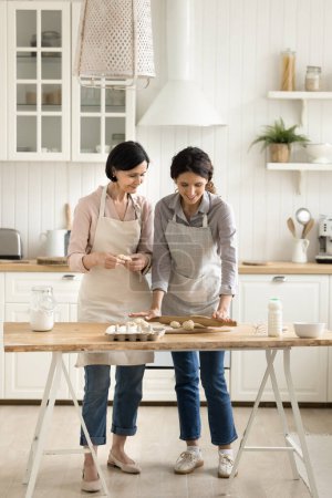 Photo for Two attractive smiling women, older mum her young adult daughter in aprons standing in kitchen kneading dough enjoy warm talk, spend weekend time at home, cooking together, prepare dinner for family - Royalty Free Image