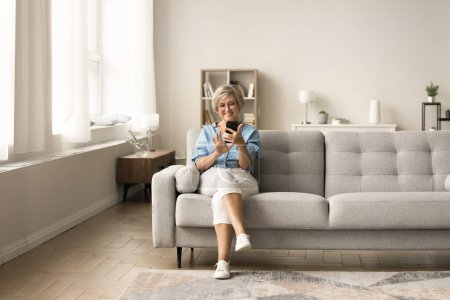 Téléchargez les photos : Heureuse femme âgée blonde élégante utilisant l'application en ligne sur téléphone mobile, en utilisant un smartphone sur le canapé, profiter des loisirs, la communication Internet dans l'intérieur de la maison moderne - en image libre de droit