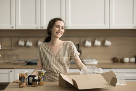 Foto de Feliz chica bastante adulta desempacando caja de cartón con verduras enlatadas en la cocina, conseguir la compra de comestibles en el supermercado, de pie en la mesa con frascos de vidrio, sonriendo, riendo - Imagen libre de derechos