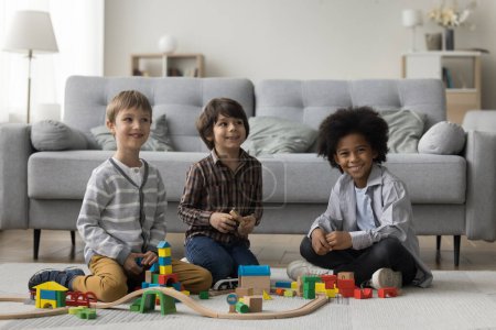 Tres niños multiétnicos felices jugando a desarrollar el juego en una casa acogedora, torre de construcción, camino en el piso cálido, mirando a la cámara, posando, sonriendo, riendo, disfrutando del ocio, tiempo de juego, actividad en el interior