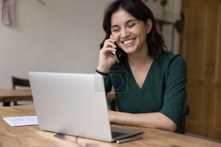 Photo for Cheerful employee woman using digital gadgets for online wireless communications in home office, sitting at work table, making business call, talking on mobile phone at laptop, smiling, laughing - Royalty Free Image