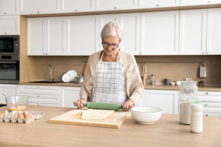 Photo for Happy blonde senior baker woman in cooking apron enjoying culinary hobby in home kitchen, baking fresh homemade pastry food, rolling dough on table with bakery ingredients, smiling - Royalty Free Image