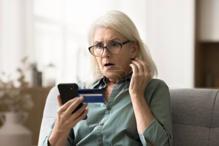 Foto de Preocupado mujer de edad avanzada sorprendida en gafas mirando el teléfono inteligente y la tarjeta de crédito, conseguir problemas financieros, mal servicio bancario en línea, la búsqueda de gasto excesivo, riesgo de quiebra - Imagen libre de derechos