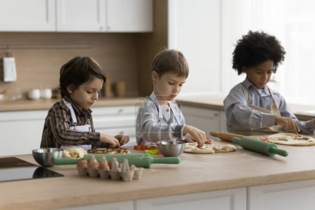 Aprendiz multiétnico enfocado niños hornear postre de pastelería en casa, dar forma, cortar masa para galletas, aprender a preparar comida casera de panadería en la mesa con utensilios, ingredientes