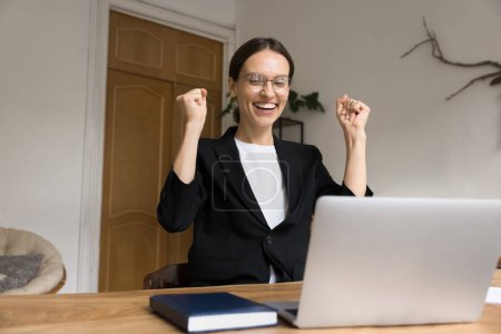 Photo for Cheerful excited winner business woman enjoying triumph, win, job achievement, success, making winner hand gesture at laptop, sitting at home office work table, laughing, celebrating profit - Royalty Free Image