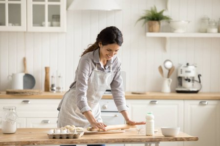 Photo for Young 35s Hispanic woman in apron standing in cozy domestic kitchen, flatten homemade dough using rolling-pin, prepare pastries for family lunch. Lifestyle, homemade food preparation, routine, chores - Royalty Free Image
