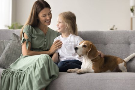 Photo for Happy pretty mom cuddling cute little daughter, stroking adorable dog on cozy home sofa, playing with sweet pet at home. Mother enjoying motherhood, family leisure with preschool girl and beagle - Royalty Free Image