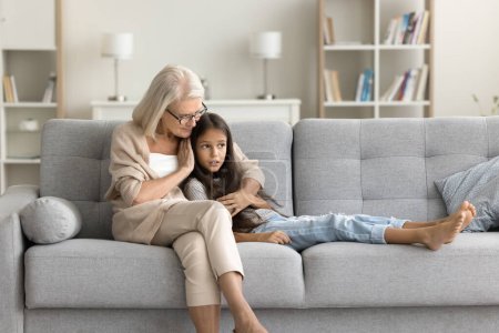 Photo for Serious grandmother and granddaughter kid talking with trust on comfortable couch at home. Caring grandma comforting grandkid, hugging girl with love, warmth, giving compassion - Royalty Free Image