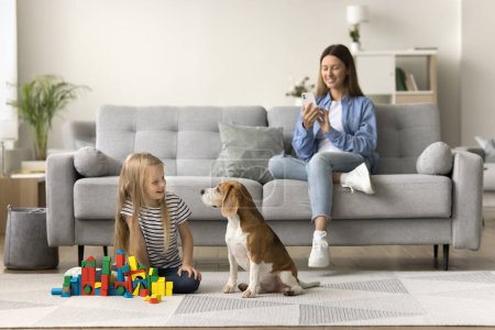 Photo for Happy mom taking picture of pet and child, sitting on comfortable sofa, watching little kid and dog playing with building cubes on warm floor, using mobile phone, shooting, recording - Royalty Free Image