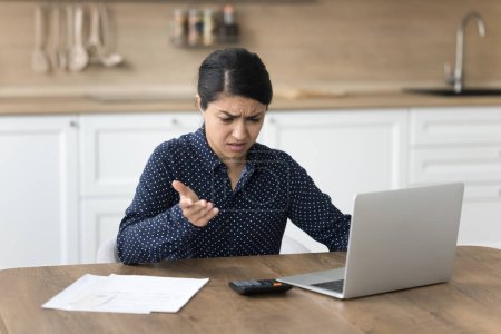 Photo for Concerned young Indian accountant woman annoyed by financial problems, bad news, finding money loss, overspending, fraud, cheating, looking at calculator at home table with laptop, documents - Royalty Free Image