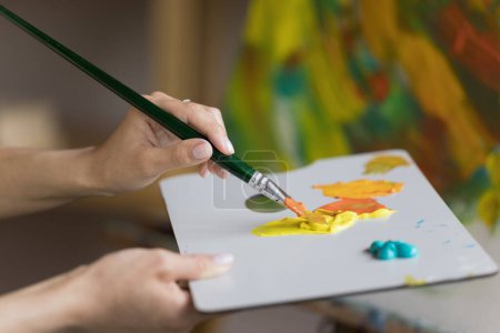 Photo for Palette of artist with acrylic paints close up. Art school student drawing picture in oil, combining yellow, orange colors on pallet, holding craft tools. Close up of hands - Royalty Free Image