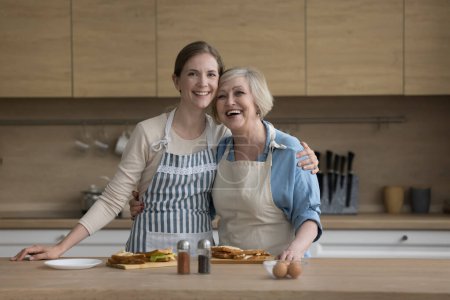 Photo for Joyful happy pretty older mom and adult daughter woman wearing aprons, standing in home kitchen at table with snacks, meal, food, hugging with love, posing for shooting, looking at camera - Royalty Free Image
