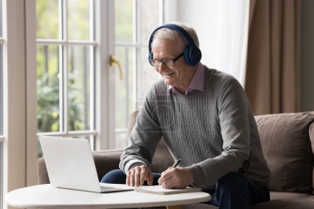 Photo for Happy older pensioner man in headphones writing notes at laptop, looking at display, speaking, laughing, using modern technologies for remote learning, Internet communications - Royalty Free Image