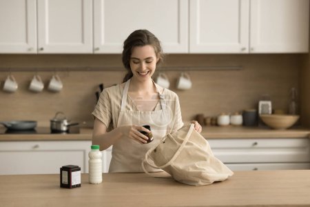 Foto de Mujer joven bonita positiva en delantal desempacar el bolso de la compra en la cocina, conseguir la compra de comestibles, botella de leche de saco de tela a la mesa, sosteniendo el frasco, sonriendo, riendo, preparando ingredientes - Imagen libre de derechos