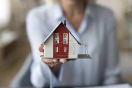 Photo for Close up female agent showing to camera small miniature of modern cottage house. Real estate agent offering new built, affordable dwelling, bank loan and property construction or remodelling services - Royalty Free Image