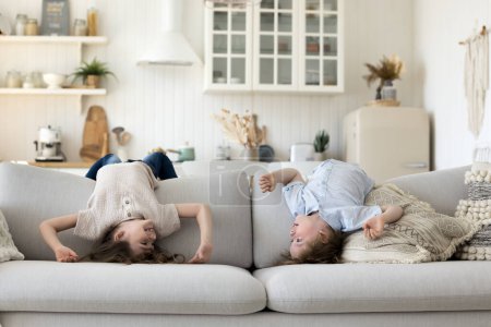 Foto de Dos lindos niños hermanos energéticos divirtiéndose en casa, disfrutando de la actividad, estirándose en el sofá, colgando boca abajo, jugando juegos activos, hablando, sonriendo, riendo - Imagen libre de derechos