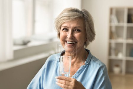 Photo for Cheerful old mature woman holding clear glass of fresh mineral water, promoting hydration for elderly skincare, healthy metabolism, healthcare, looking at camera with beautiful toothy smile - Royalty Free Image