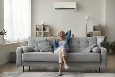 Pretty aged woman relaxing on sofa holding remote control managing temperature inside modern living room, Cooling space, make environment more comfortable at hot weather, reduce humidity level indoor