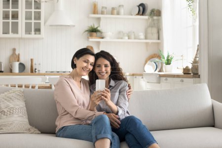 Photo for Happy mature mother hugging her millennial daughter using smart phone at home, sitting on couch staring at gadget screen make videocall to family, watch photos, read news, enjoy new mobile application - Royalty Free Image
