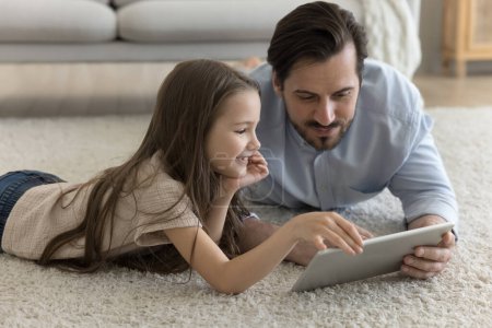 Téléchargez les photos : Un père millénaire positif et une petite fille enfant utilisant une tablette numérique d'application d'apprentissage, reposant sur un sol doux et chaud à la maison, souriant. Père et enfant jouissant de loisirs, communication, regarder un film - en image libre de droit