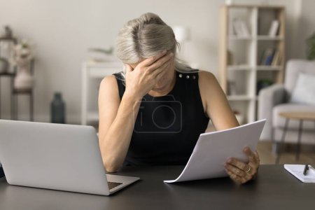 Photo for Depressed sad mature entrepreneur, business woman reading paper document at home, sitting at work table with laptop, learning bad shocking news, covering face, getting problems, stress - Royalty Free Image
