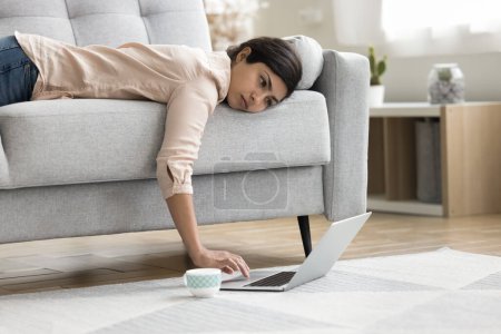 Photo for Overworked exhausted Indian freelancer woman lying on sofa with face on seat, using laptop computer placed with cup of coffee on floor, having problems with deadline, feeling tired, fatigue - Royalty Free Image