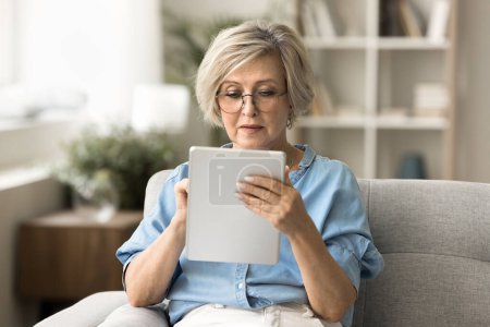 Photo for Focused blonde senior woman in glasses browsing Internet on tablet, using digital gadget, wireless technology at home, consulting doctor with medical healthcare online application - Royalty Free Image