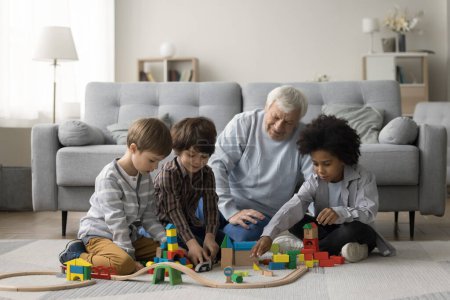 Abuelo anciano viendo a tres nietos multiétnicos jugando con bloques de construcción de juguetes en el piso cálido, construyendo torres de la ciudad modelo juntos, disfrutando del tiempo de juego familiar, trabajo en equipo