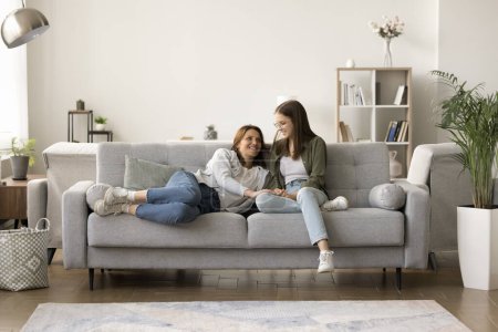 Photo for Positive mother and teenage kid resting on comfortable soft couch, relaxing in modern apartment interior, enjoying leisure in cozy home interior, talking, chatting, laughing. Full length - Royalty Free Image