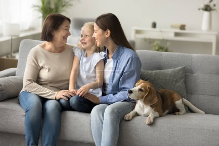 Foto de Madre feliz, abuela y niña pequeña sentada en el sofá en el adorable perro beagle, hablando, riendo, disfrutando de la reunión familiar, ocio hogareño, vinculación, afecto, sonriendo, riendo - Imagen libre de derechos