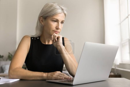 Foto de Mujer freelancer madura reflexiva que trabaja en el ordenador portátil en casa, mirando a la pantalla con cara reflexiva, tocando la barbilla, pensando, leyendo, viendo contenido en línea, tomando decisiones en pensamientos profundos - Imagen libre de derechos