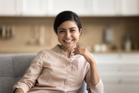 Foto de Feliz hermosa mujer india joven mirando a la cámara con sonrisa dentada, sentado en el sofá en casa, riendo, hablando en videollamada informal en línea, disfrutando del ocio, comodidad. Retrato en la cabeza - Imagen libre de derechos