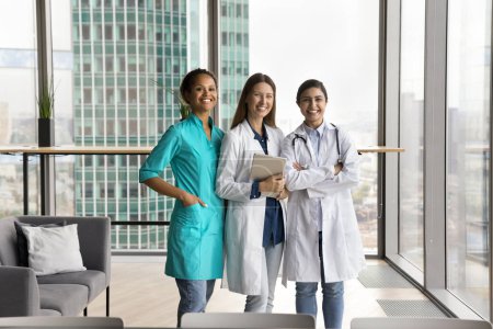 Téléchargez les photos : Heureuse jeune femme médecin multiethnique confiante en uniforme debout ensemble dans la salle de bureau de l'hôpital, regardant la caméra, souriant, riant. Équipe diversifiée de femmes médecins portrait - en image libre de droit