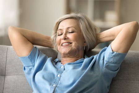 Foto de Mujer jubilada con calma positiva descansando en un cómodo sofá en casa, que conduce al sofá con los codos abiertos, relajándose con los ojos cerrados y la sonrisa dentada, respirando aire fresco - Imagen libre de derechos