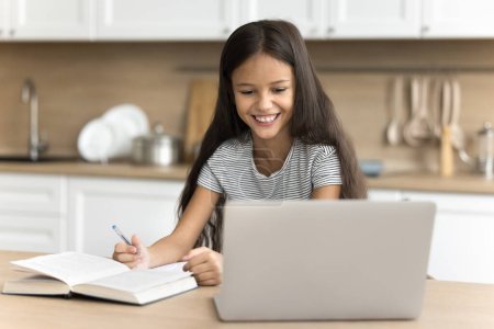Foto de Feliz niña preadolescente estudiando en línea en casa, haciendo tarea escolar, sentada en la mesa y aprendiendo libro, escribiendo notas, mirando la pantalla, viendo la lección en Internet, sonriendo - Imagen libre de derechos