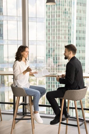 Photo for Two young entrepreneurs discussing cooperation in co-working space, talking at window with urban downtown view background, smiling, enjoying networking, collaborating on project - Royalty Free Image