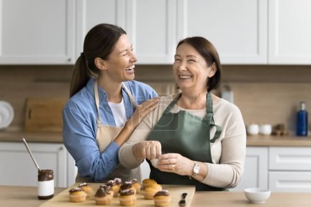 Téléchargez les photos : Joyeuse jeune fille adulte excitée femme aidant maman aînée à décorer de délicieux cupcakes au chocolat, embrassant les épaules des mamans, parlant, riant, appréciant les activités de cuisine familiale - en image libre de droit