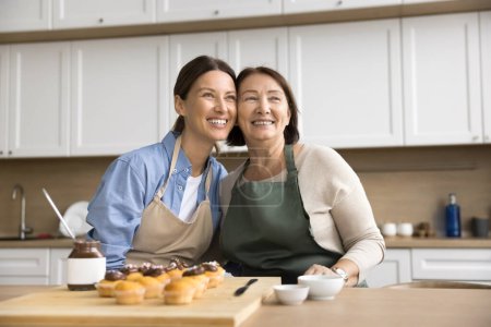 Téléchargez les photos : Joyeux jeune fille adulte et maman à la retraite dans des tabliers préparant des muffins sucrés faits maison pour le dessert, étreignant à table avec des cupcakes au chocolat, regardant loin avec des sourires rêveurs - en image libre de droit