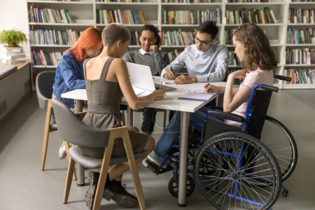 Photo for Diverse group of students inclusively college mate with disability sitting at table in library together, writing notes, working on creative essay, doing school homework together. Full length - Royalty Free Image