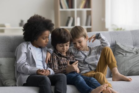 Equipo multiétnico de niños pequeños enfocados que usan el teléfono móvil juntos, descansando en un acogedor sofá en casa. Niño preescolar jugando videojuego en línea en el teléfono inteligente, mostrando la pantalla a los amigos