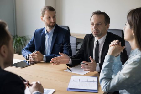 Photo for Serious business man in formal suit talking to partners on meeting, discussing deal, partnership, investment, project strategy. Team leader, CEO, executive speaking to colleagues, employees at table - Royalty Free Image