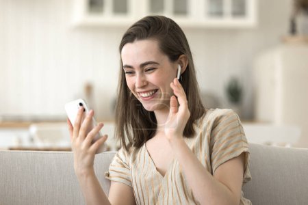 Photo for Positive young adult girl talking on video call on cellphone, sitting on couch, holding smartphone, using wireless earphones, speaking at screen, laughing, waving greeting hand hello - Royalty Free Image
