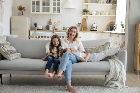 Photo for Positive young mom enjoying motherhood, leisure time at home with sweet little son and daughter, resting on soft couch with children, talking to kids, smiling, laughing - Royalty Free Image