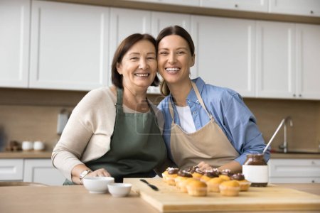 Téléchargez les photos : Bonne maman aînée et adulte enfant femme dans des tabliers de boulanger préparant le dessert maison pour la fête de famille, debout à table avec des cupcakes au chocolat, étreignant, regardant la caméra, souriant pour le portrait - en image libre de droit