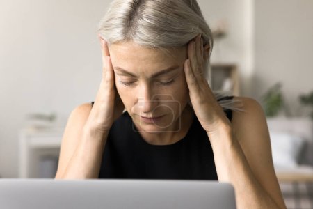 Téléchargez les photos : Femme blonde mature fatiguée dans la tête touchante occasionnelle à l'ordinateur portable, assise à la table de travail, fermant les yeux, souffrant de maux de tête, migraine, se sentant stressée, choquée, surmenée, malade - en image libre de droit